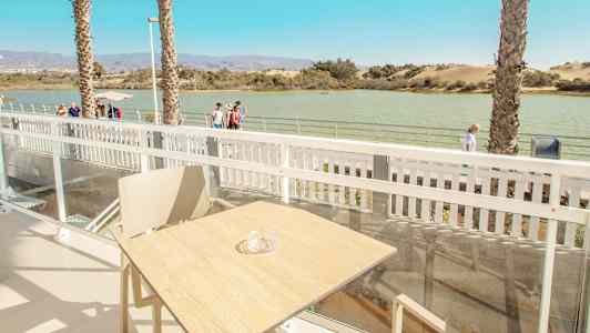 Utsikt fra en terrasse med bord og stol, med vann og gangvei i bakgrunnen. (AI-generert tekst)