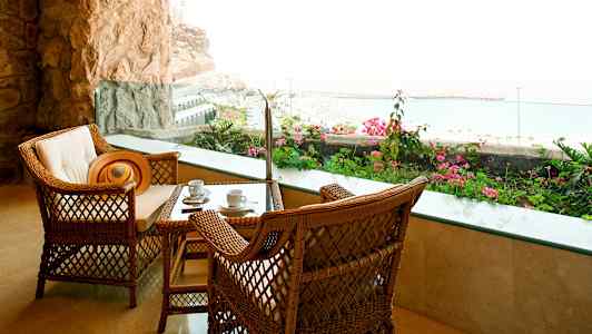 To rottingstoler og et bord med kaffekopper, på en terrasse med utsikt over havet og blomsterprakt. (AI-generert tekst)