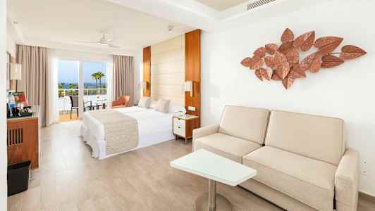 Lyst hotellrom med seng, sofa og balkong samt havutsikt. (AI-generert tekst)