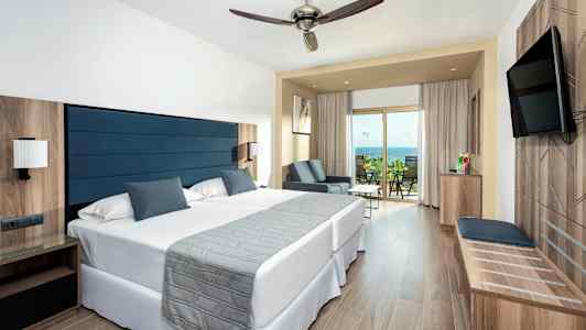 Et hotellrom med to senger, en sitteplass og en balkong med utsikt over havet. (AI-generert tekst)