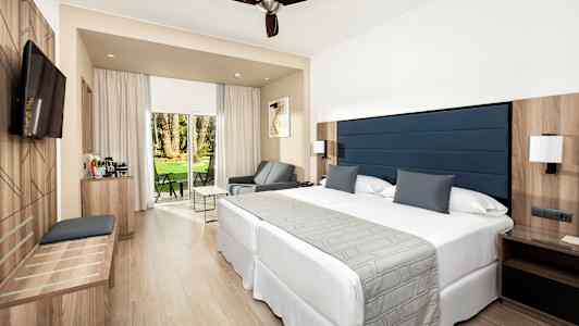 Et moderne hotellrom med en stor seng, sittemøbler og en glassfront med utsikt til en frodig hage. (AI-generert tekst)