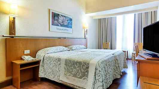 Et hotellrom med en dobbeltseng med mønstret overkast, lamper på veggen og en TV på et skrivebord. Sitteplasser og et vindu med gardiner er også til stede. (AI-generert tekst)