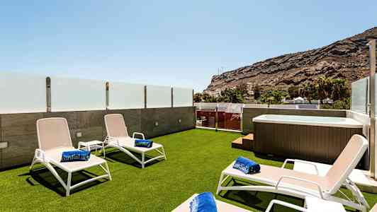 Terrasse med solsenger, badehåndklær og boblebad, omgitt av glassrekkverk og utsikt mot fjell. (AI-generert tekst)