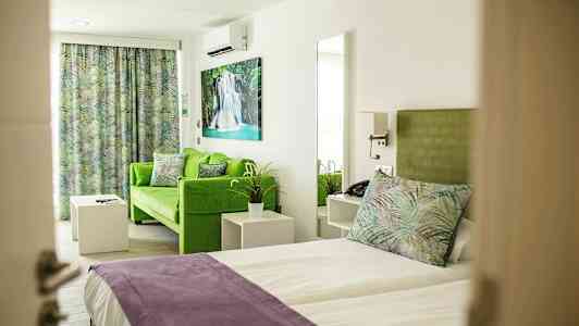 Rom med seng og grønn sofa, med fargerike puter og gardiner samt kunst på veggen. (AI-generert tekst)