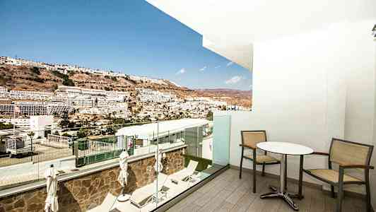 Utsikt fra en balkong med bord og stoler ut mot en åsside med bygninger i det fjerne. (AI-generert tekst)