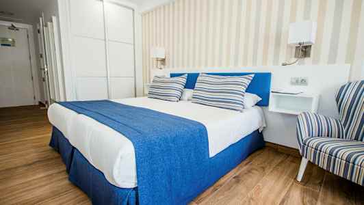 Et elegant hotellrom med en dobbeltseng dekket med blått og hvitt sengetøy samt stripete puter, ved siden av en stripete lenestol. (AI-generert tekst)