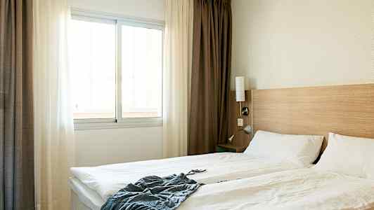 Et hotellrom med en dobbeltseng, hvite sengeklær, en trepanel ved sengegavlen og et vindu med lyse gardiner. (AI-generert tekst)