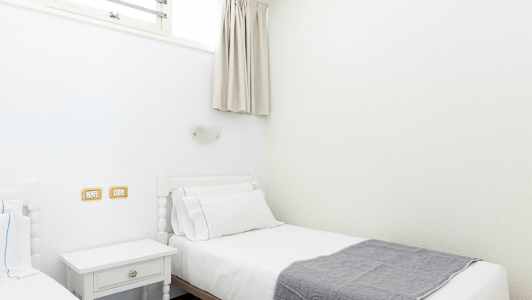 To enkeltsenger med hvitt sengetøy og et grått teppe, plassert i et lyst rom. (AI-generert tekst)