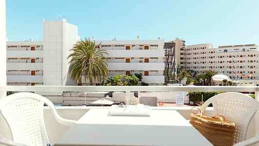 Utsikt fra en balkong med sittegruppe, bygninger og palmer synlige i bakgrunnen under en klar blå himmel. (AI-generert tekst)