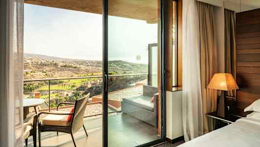 Et hotellrom med utsikt gjennom en glassdør mot en balkong med stoler og et fjernt landskap utendørs. (AI-generert tekst)