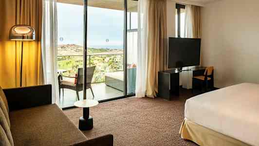 Hotellrom med sittegruppe, seng, TV og utsikt mot en balkong med landskap utenfor. (AI-generert tekst)