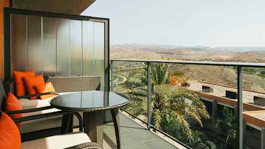 Balkong med sittemøbler, glassbord og utsikt over omgivelser med palmer og landskap. (AI-generert tekst)