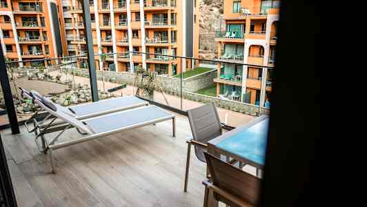 To solsenger og et bord med stoler på en romslig balkong med utsikt over en gårdsplass og flere oransje bygninger. (AI-generert tekst)