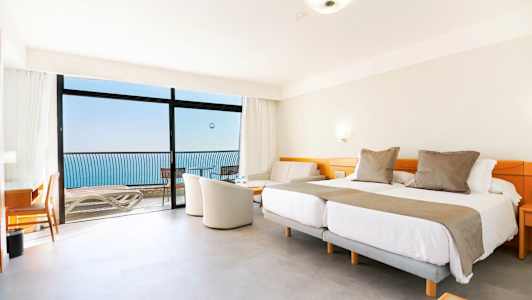 Et hotellrom med dobbeltsenger, sittegruppe og balkong med havutsikt. (AI-generert tekst)