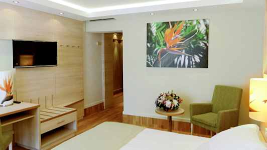 Et hotellrom med en seng, grønn stol, blomsterbukett på et bord og et veggbilde av en oransje blomst. (AI-generert tekst)