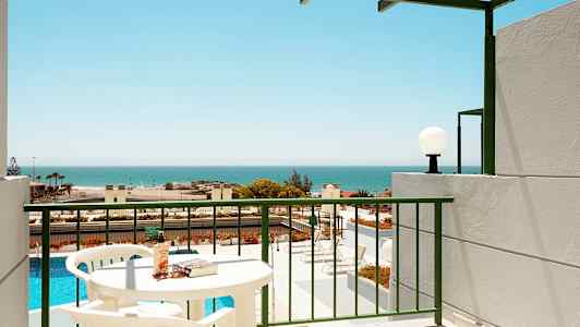 En balkong med utsikt over et basseng og havet, bord og stoler synlige. (AI-generert tekst)