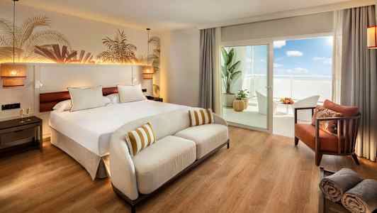 Et hotellrom med en stor seng, en hvit sofa og en balkong med møbler. (AI-generert tekst)