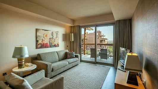 En stue med en sofa, lenestol og et salongbord, en TV på et møbel samt en glassdør som fører ut til en balkong. (AI-generert tekst)