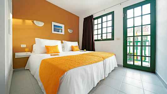 Et soverom med en dobbeltseng med hvite og oransje tekstiler, og store vinduer med grønne karmer. (AI-generert tekst)