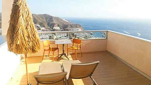 Utsikt fra en terrasse med parasoll, to stoler og et bord, samt solsenger med havutsikt. (AI-generert tekst)