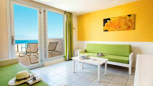 En lys stue med en grønn sofa, et hvitt salongbord og utgang til balkong med havutsikt. (AI-generert tekst)