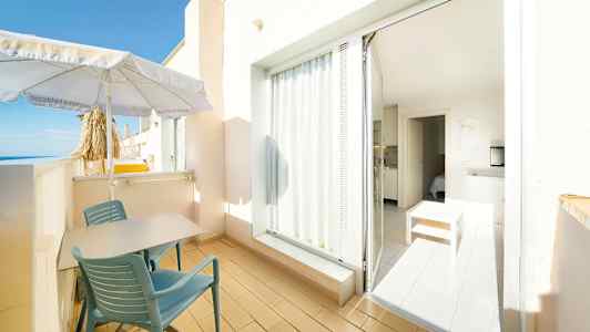 En balkong med bord og stoler, parasoll og utsikt mot havet. En dør leder til et lyst interiør med møbler. (AI-generert tekst)