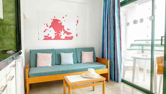 En stue med en turkis sofa, fargerike puter, bord og en hatt. Et vindu med utsikt mot en balkong. (AI-generert tekst)