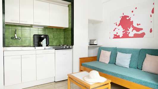 Et enkelt kjøkken med hvit innredning og grønne fliser bak. Ved siden av er det en sofa med turkise puter og pastellfargede puter. (AI-generert tekst)