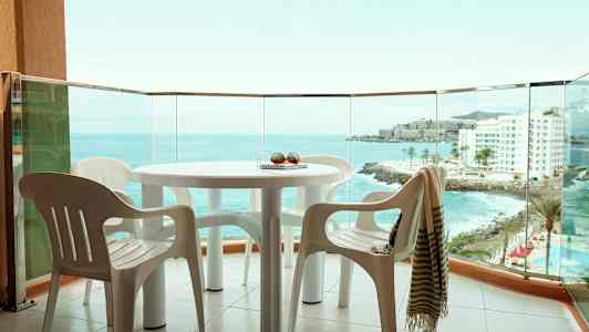 Utsikt fra en balkong med et bord og plaststoler, havet og bygninger synlige i bakgrunnen. (AI-generert tekst)