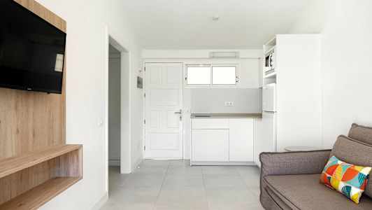 Et hvitt rom med en sofa, TV, trehylle og kjøkken. (AI-generert tekst)