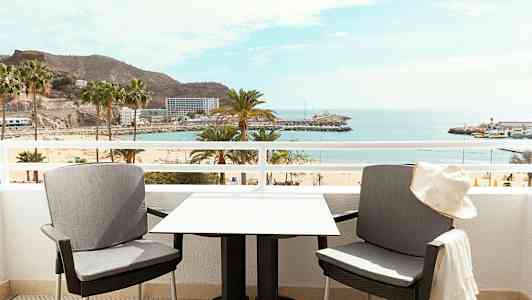 To stoler og et bord på en balkong med utsikt over en strand og havet. (AI-generert tekst)