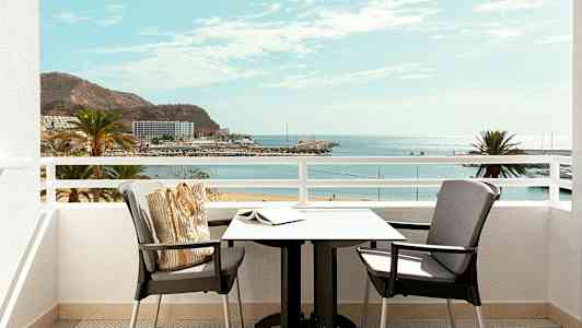 Utsikt fra en balkong med bord og stoler mot en strand og havet. (AI-generert tekst)