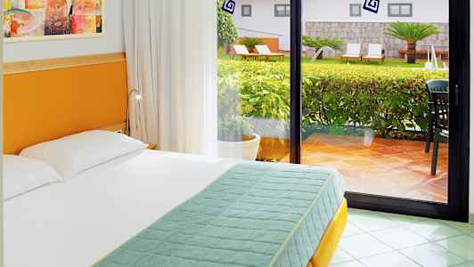 Et hotellrom med en seng foran et stort glassvindu som viser utsikt over en frodig hage. (AI-generert tekst)