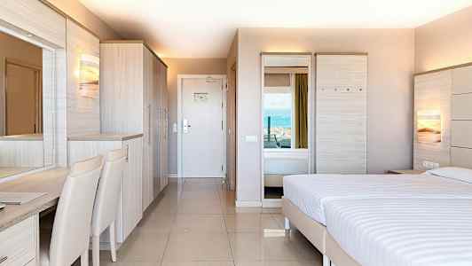 Et hotellrom med moderne innredning, en dobbeltseng, garderobe og skrivebord, med utsikt mot havet. (AI-generert tekst)