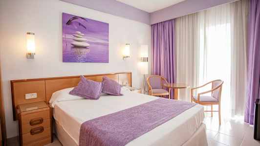 Hotellrom med dobbeltseng, lilla detaljer og sitteplass ved et vindu. (AI-generert tekst)