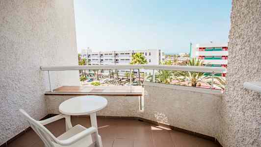 Bilde av en balkong med bord og stol, med utsikt over bygninger og palmer. (AI-generert tekst)