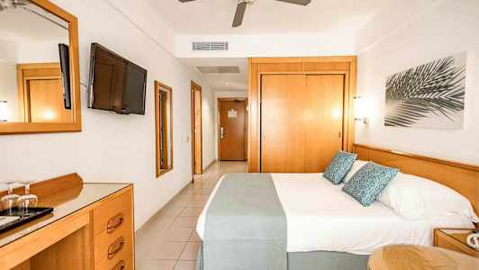 Et hotellrom med en dobbeltseng, TV på veggen, takvifte og tremøbler. (AI-generert tekst)