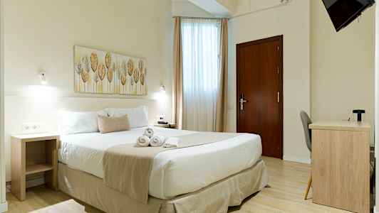 Et lyst hotellrom med en dobbeltseng, et skrivebord og en veggmontert TV. (AI-generert tekst)