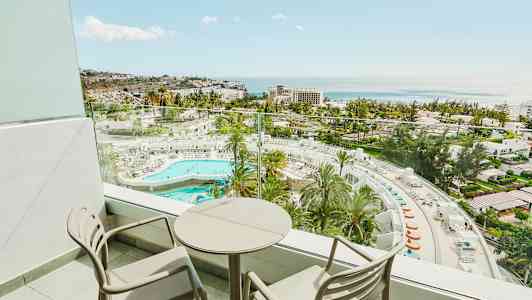 Balkong med to stoler og et rundt bord samt utsikt over hotellområde med basseng og hav. (AI-generert tekst)