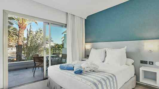 Soverom med dobbeltseng, hvitt sengetøy og blå dekorativ vegg, samt balkong med stoler og utsikt over palmer. (AI-generert tekst)