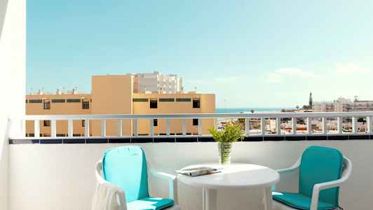 En balkong med to stoler, et bord og utsikt over bygninger og havet. (AI-generert tekst)