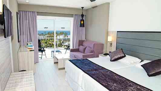 Moderne hotellrom med en dobbeltseng, sofagruppe og balkong med utsikt over havet. (AI-generert tekst)