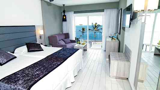 Hotellrom med dobbeltseng, sofa og havutsikt utenfor store vinduer. (AI-generert tekst)