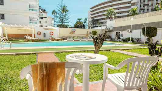 Et utendørs basseng omgitt av grøntområder og solsenger, med hvite bygninger i bakgrunnen. (AI-generert tekst)