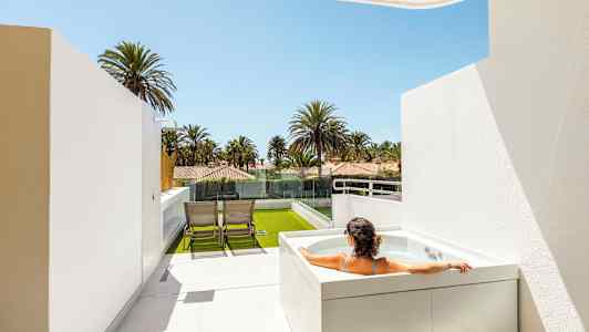 En person sitter i et boblebad på en terrasse med utsikt over palmer og hus. (AI-generert tekst)