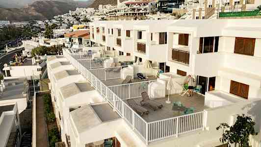 Bilde av en rekke moderne hvite leiligheter med store private balkonger, møblert med stoler og solsenger, i en kupert omgivelser med fjell i bakgrunnen. (AI-generert tekst)