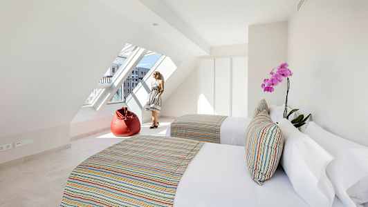 Et lyst rom med to senger, fargerike puter, en rød sittesekk og et stort skråstilt vindu. (AI-generert tekst)