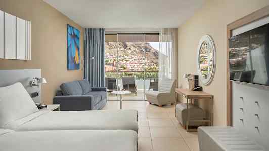 Hotellrom med to senger, sofa, lenestol og stor balkong med utsikt mot et fjellagt nabolag. (AI-generert tekst)