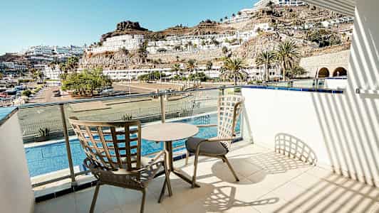 En balkong med to stoler og et bord, utsikt over et basseng, bygninger og fjell. (AI-generert tekst)