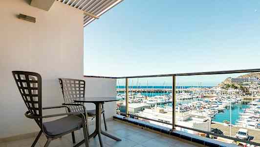 Bildet viser en balkong med to stoler og et bord, med utsikt over en havn med båter. (AI-generert tekst)